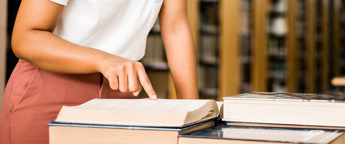 Person pointing to text in an open book while standing in a library.